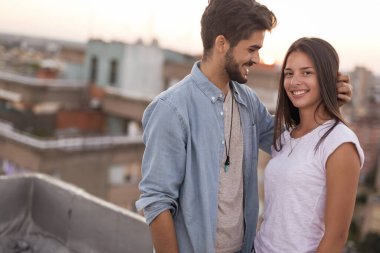 Aşık bir çift gün batımında bir binanın çatısında dikiliyor ve sarılıyor. Arka planda şehir manzarası var.