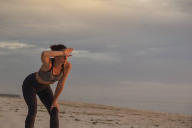 Yoğun bir egzersizden sonra sahilde duran genç bir kadın, sabahın erken saatlerinde sahilde koştuktan sonra dizlerinin üstüne çöküp ter siliyor.