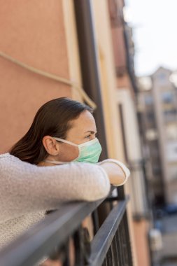 Koronavirüs koruma ve önleme protokollerinin bir parçası olarak dışarıda cerrahi maske takan, balkonda tek başına duran genç bir kadının portresi.
