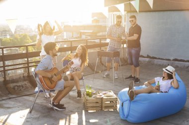 Bir grup genç arkadaş çatıdaki partide eğleniyor, barbekü yapıyor, bira içiyor, gitar çalıyor ve sıcak yaz günlerinin tadını çıkarıyor. Sandalyede oturan kıza odaklan.