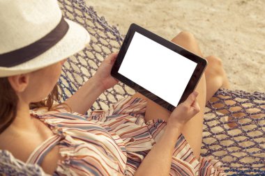 Kumsalda bir hamakta oturmuş, elinde boş ekranlı bir tablet bilgisayar tutan bir kadın.
