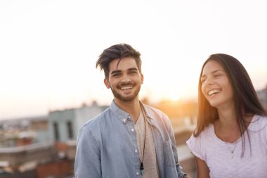 Aşık bir çift gün batımında bir binanın çatısında dikiliyor ve sarılıyor. Arka planda şehir manzarası var.