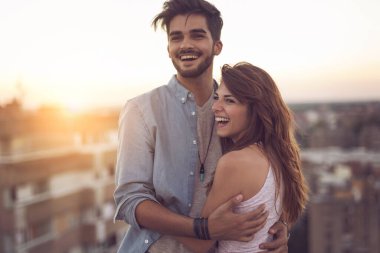 Aşık bir çift gün batımında bir binanın çatısında dikilip sarılıyor ve arka planda şehir manzarası var. Kıza odaklan.