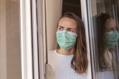 Hasta kadın dairesinin içinde dikilip pencereden dışarı bakıyor, tıbbi yüz koruma maskesi takıyor, kovid-19 virüsü salgını sırasında evde yalnızlık çekiyor.