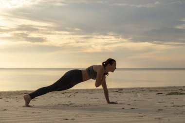 Sabah plajda egzersiz yapan, omuz vuruşları yapan genç bir kadının yan görüntüsü.