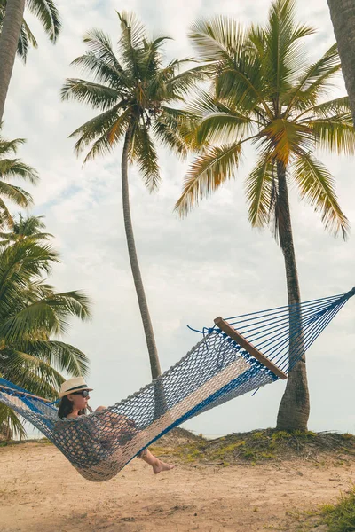 Genç bir kadın tatildeyken dinleniyor, hamakta oturuyor palmiye ağaçlarının arasına bağlanmış güzel tropikal plajda.