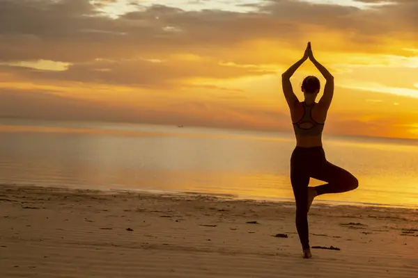 stock image Active young woman doing yoga on the beach, holding a tree pose or Vrikshasana, standing balancing yoga pose and enjoying beautiful sunrise over the water