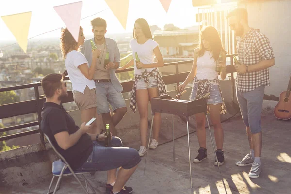 Group of young friends having fun at rooftop party, making barbecue, drinking beer and enjoying hot summer days. Focus on the people next to the fence