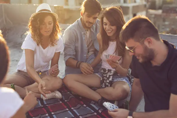 ピクニックブランケットに座っている若者のグループは 建物の屋上でカードをプレイしながら楽しんでいます 右の女の子に焦点を当てる — ストック写真
