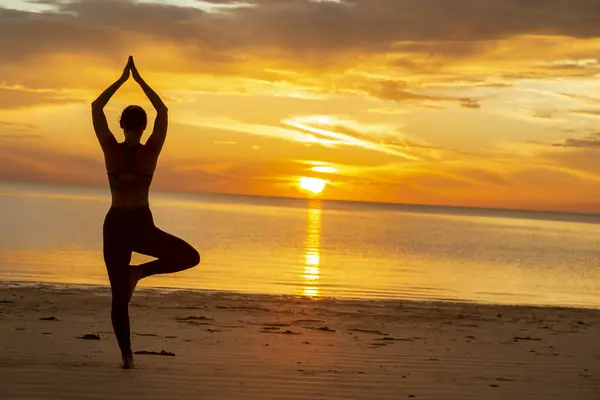 stock image Active young woman doing yoga on the beach, holding a tree pose or Vrikshasana, standing balancing yoga pose and enjoying beautiful sunrise over the water