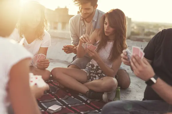 Group of young people sitting on a picnic blanket, having fun while playing cards on the building rooftop. Focus on the couple in the middle