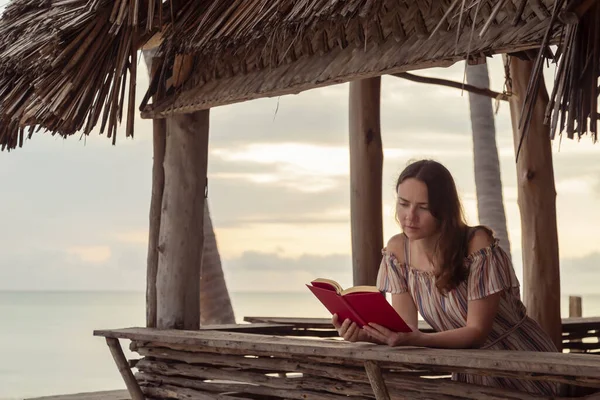 エキゾチックな熱帯ビーチバーカウンターに立って 本を読み 休暇中に海の上に美しい夕日を楽しむ若い女性 — ストック写真
