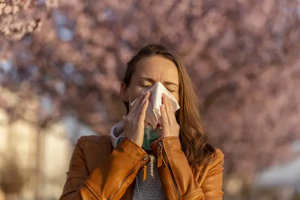 Portrait of a young woman sneezing and blowing nose outdoor, having trouble with spring pollen allergies and inhaler allergens