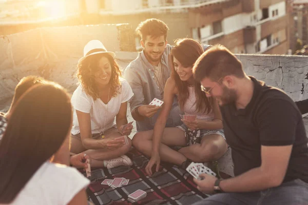 Group of young people sitting on a picnic blanket, having fun while playing cards on the building rooftop. Focus on the guy on the right