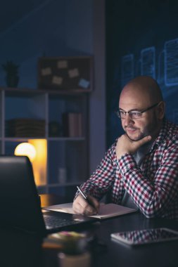 Fazla mesai yapan bir adam masasında oturup online bir toplantı yapıyor, evden çalışırken bir planlamacıdan not alıyor.