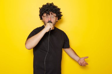 Peruk takan adam portresi, siyah tişört ve gözlük takan mikrofon taşıyan ve sarı arka planda izole edilmiş şarkı söyleyen fotokopi çeken.