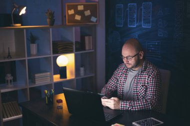 Ofisteki masasında oturan bir adam, gece geç saatlere kadar çalışırken akıllı telefon kullanarak mesaj yazıyor.
