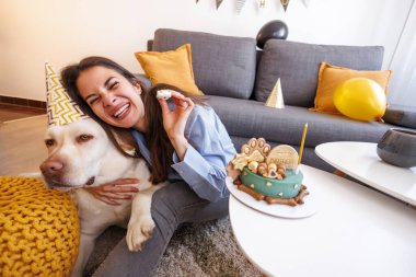 Güzel, neşeli genç bir kadın labrador köpeğinin doğum gününü evde kutluyor, parti şapkası takıyor ve parti düdüğü çalıyor.