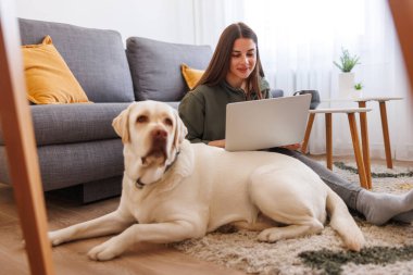 Evinde bilgisayarla uzaktan çalışırken köpeğine sarılan güzel bir genç kadın. Evcil hayvanıyla evde çalışan serbest çalışan bir kadın.