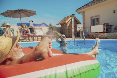 Bir grup yaşlı insan sıcak yaz günlerini yüzme havuzunda geçiriyorlar, güneşleniyorlar, şişme yatakta yüzüyorlar ve birbirlerine su sıçratıyorlar, serinliyorlar ve dinleniyorlar.