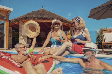 Güneşli bir yaz gününde, yüzme havuzunda güneşlenen ve eğlenen iki yaşlı çift tatillerinin tadını çıkarıyor, kokteyller içiyor ve havuzda serinliyorlar.