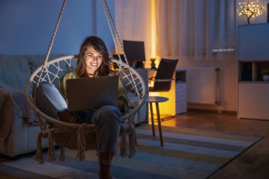 Evde boş vakitten zevk alan bir kadın, oturma odasında sallanan bir sandalyede oturuyor, dizüstü bilgisayar kullanıyor, arkadaşlarıyla sohbet ediyor ve gece geç saatlerde dinleniyor.