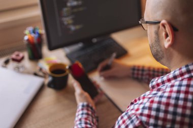 Akıllı telefonu tutan ve evden çalışırken not alan programcının yüksek açılı görüntüsü