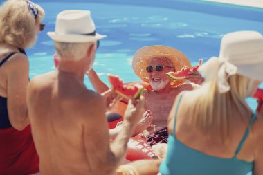 Bir grup neşeli son sınıf öğrencisi yüzme havuzunun kenarında oturup karpuz yiyor, güneşleniyor ve sıcak bir yaz gününde dışarıda eğleniyorlar.