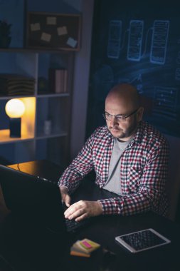 Erkek ağ geliştirici masasında oturup ev ofisinde fazla mesai yapıyor, dizüstü bilgisayar kullanıyor.