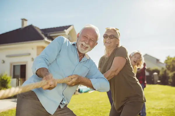 Senior people having fun playing tug of war, spending sunny summer day outdoors; group of elderly friends having fun participating in rope pulling competition at retirement home