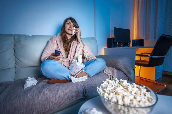 Beautiful young woman sitting on couch covered with blanket late at night, sad while watching drama movie on TV, crying and wiping tears with paper tissues