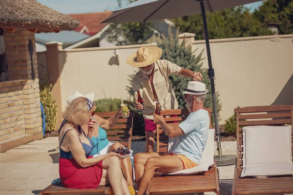 Group of elderly friends relaxing and sunbathing on sun beds by the swimming pool while on a summer vacation, making a toast, drinking cocktails and beer and having fun