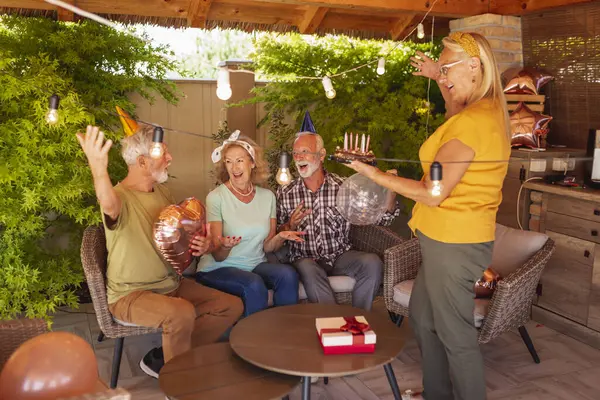Group of cheerful senior friends throwing a surprise birthday party for a friend, wearing party hats, holding balloons and singing birthday song while bringing cake
