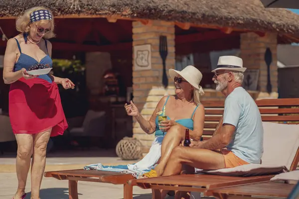 Group of elderly friends having fun while on a summer vacation, sitting on sunbeds by the swimming pool, drinking cocktails, eating fresh fruit and relaxing