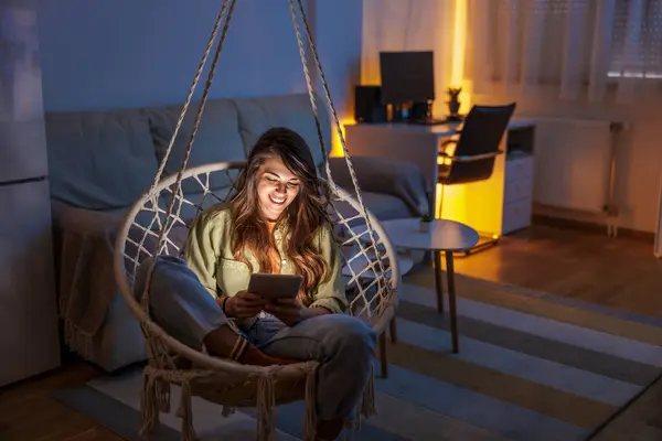 美しい若い女性は 電子ブックを読んで吊るす椅子に座って自宅で余暇を楽しんでいます — ストック写真
