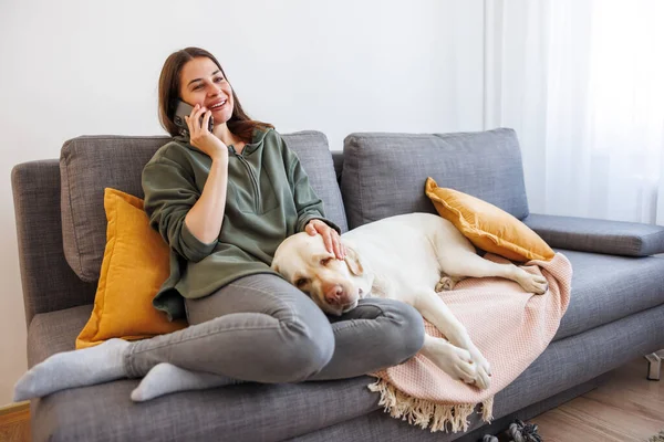 快乐的年轻女人在家里休息和享受闲暇时光 打电话 抱着宠物狗躺在腿上 — 图库照片