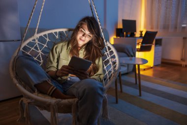 Güzel genç bir kadın sallanan sandalyede oturmuş e-kitap okuyor e-okuyucu kullanıyor ve evde dinleniyor.