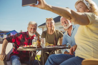 Bir grup neşeli son sınıf öğrencisi havuz kenarında öğle yemeği yerken selfie çekiyor, masanın etrafında toplanıyor, güneşli bir yaz gününde yiyor, içiyor ve dinleniyorlar. 