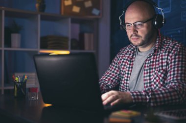 Kulaklık takan adam gece geç saatlerde ev ofisinde dizüstü bilgisayarla çalışıyor; web geliştiricisi fazla mesai yapıyor