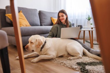 Evinde bilgisayarla uzaktan çalışırken köpeğine sarılan güzel bir genç kadın. Evcil hayvanıyla evde çalışan serbest çalışan bir kadın.