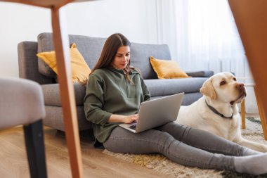Güzel, genç bir kadın köpeğiyle birlikte oturma odasında oturuyor. Uzaktan bilgisayarla evden çalışıyor.