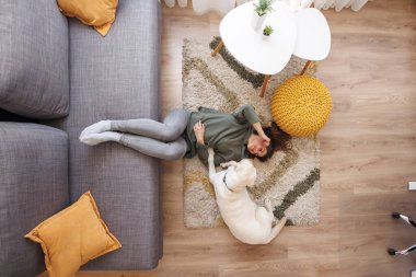 Genç ve güzel bir kadının evde dinlenip evcil köpeği Labrador 'la vakit geçirmesinin manzarası. İkisi de oturma odasında yerde yatıyor.