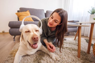 Güzel genç bir kadın evde köpeğiyle vakit geçiriyor, kulaklık takıyor ve müzik dinliyor.