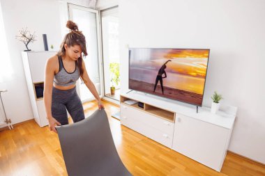 Spor giyim sektöründe aktif genç bir kadın. Oturma odasının zeminine yoga minderi döşeyerek online ev yapımı yoga seansına hazırlanıyor.