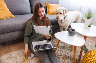 Güzel genç bir kadın uzaktan bilgisayarla çalışıyor ve evcil köpeği yanında dururken evrak işleriyle uğraşıyor.