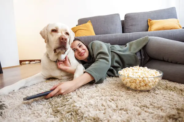 Beautiful young woman cuddling her pet dog while relaxing and enjoying leisure time at home, lying on living room floor eating popcorn and watching TV