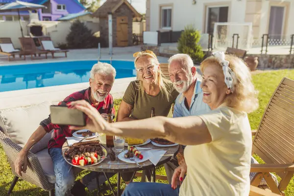 Group of elderly friends taking a selfie while having lunch in the backyard by the pool, gathered around the table, eating, drinking and enjoying sunny summer day outdoors