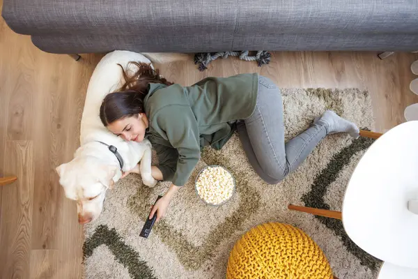 Top view of beautiful young woman cuddling her pet dog while relaxing and enjoying leisure time at home, lying on living room floor eating popcorn and watching TV
