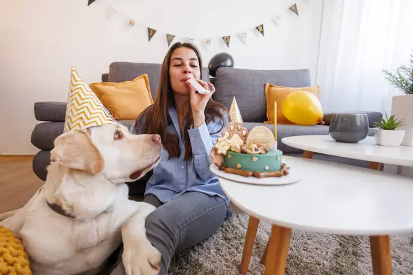 Beautiful cheerful young woman having fun celebrating her pet labrador dog\'s birthday at home, wearing party hats and blowing party whistles