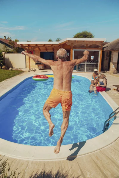 Group of elderly friends relaxing and sunbathing by the swimming pool on a hot summer day, one man having fun jumping into the pool while on a vacation